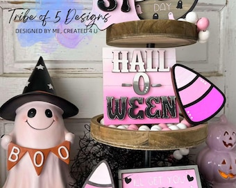 以下が含まれることがあります： ハロウィンをテーマにした装飾が施された3段の木製トレイ。最上段には、数字の31が書かれたピンクの幽霊と、「ハッピーブーデー」と書かれた丸い看板があります。中段には、「ハローウィン」と書かれたピンクと白の看板があります。最下段には、ピンクと白のキャンディーコーン、ピンクの看板に「アイ・ゲット・ユー・マイ・プリティ」と書かれており、ピンクの幽霊に「ブー」と書かれたバナーがあります。