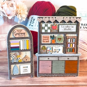 May include: A wooden craft project depicting a sewing room with a shelf, a cabinet, and various sewing-themed decorations. The shelf has fabric scraps, yarn, and a sewing iron. The cabinet has drawers and a sewing machine. The decorations include a sign that says "Welcome to my Sewing Room", a sign that says "I'd rather be sewing", and a sign that says "Sewing mends the soul".