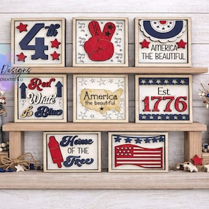 May include: A collection of patriotic wooden signs with red, white, and blue designs. The signs feature phrases like "4th", "America the Beautiful", and "Home of the Free", along with star and flag motifs. Decorative elements include star-shaped accents.