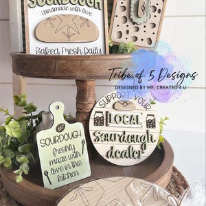 May include: A set of five wooden signs with a sourdough bread theme. The signs feature phrases like "Sourdough Handmade with Love", "Baked Fresh Daily", "Support Your Local Sourdough Dealer", and "Sourdough Freshly Made with Love in This Kitchen".