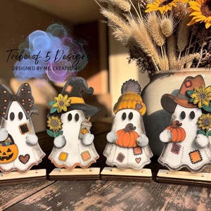 May include: Four white ghost figurines standing on wooden bases. The first wears a black bow and holds a jack-o'-lantern bucket. The second wears a witch hat with sunflowers and holds a drink. The third wears a beanie and holds a pumpkin. The fourth wears a cowboy hat with sunflowers and holds a pumpkin.