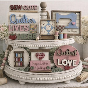 Peut inclure: Présentation sur un plateau à plusieurs niveaux d'objets décoratifs sur le thème du patchwork. Panneaux en bois avec des phrases comme « Sew Cute » et « Quilted with Love ». Palette de couleurs pastel : rose, bleu et vert, avec des bobines de fil et des tissus.
