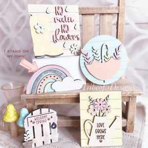 May include: A collection of pastel-colored wooden spring-themed decorations. Includes a rainbow, umbrella, watering can with flowers, a boot, and a welcome sign. The text on the decorations reads "No rain no flowers" and "Love grows here".