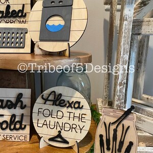 May include: Wooden laundry room decor. A round sign displays a washing machine graphic with a blue water design. Other signs include the text "Alexa Fold the Laundry" and a mason jar with clothespins. The signs are on wooden stands.