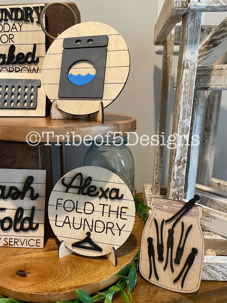 May include: Wooden laundry room decor. A round sign displays a washing machine graphic with a blue water design. Other signs include the text "Alexa Fold the Laundry" and a mason jar with clothespins. The signs are on wooden stands.