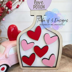 Puede incluir: Botella de poción de amor de madera con un corazón morado en la parte superior que dice "love potion". La botella tiene un fondo blanco y está decorada con corazones rojos y rosas. La botella está sobre una superficie de madera.