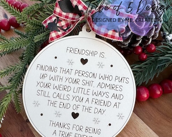 May include: White wooden ornament with a red and black checkered bow. The ornament has the text "Friendship is... Finding that person who puts up with your shit, admires your weird little ways and still calls you a friend at the end of the day. Thanks for being a true friend."