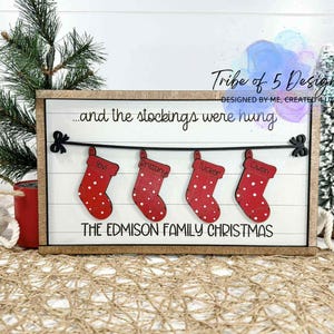 May include: A wooden Christmas sign with the text "...and the stockings were hung" and "THE EDMISON FAMILY CHRISTMAS." Four red stockings with white polka dots are displayed, each with a name. The sign is framed in a light brown wood.