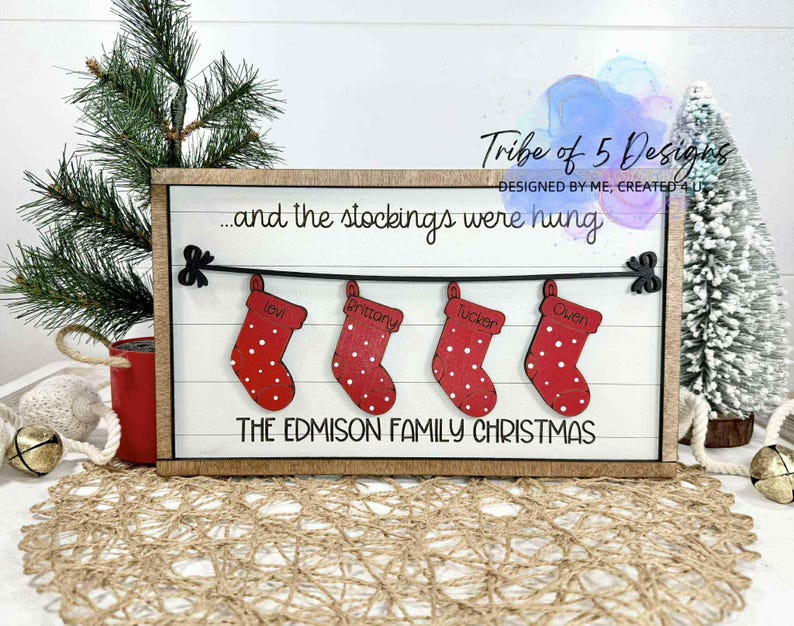 May include: A wooden Christmas sign with the text "...and the stockings were hung" and "THE EDMISON FAMILY CHRISTMAS." Four red stockings with white polka dots are displayed, each with a name. The sign is framed in a light brown wood.