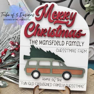 Puede incluir: Un letrero navideño de madera con las palabras "Merry Christmas" en letras rojas. El letrero presenta un coche familiar verde y marrón que transporta un árbol de Navidad, con el texto "The Mansfield Family Christmas Farm". El letrero dice "Home of the Fun Old Fashioned Family Christmas".