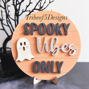 May include: A round wooden sign with a peach background and black and silver lettering. The sign reads "Spooky Vibes Only" with a white ghost illustration. The sign is on a small wooden stand.