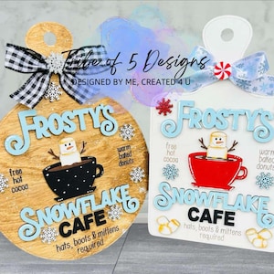 May include: Two wooden signs with a winter theme. Both signs are round and feature the text "Frosty's Snowflake Cafe" with a snowman and a cup of hot cocoa. One sign is brown and the other is white.