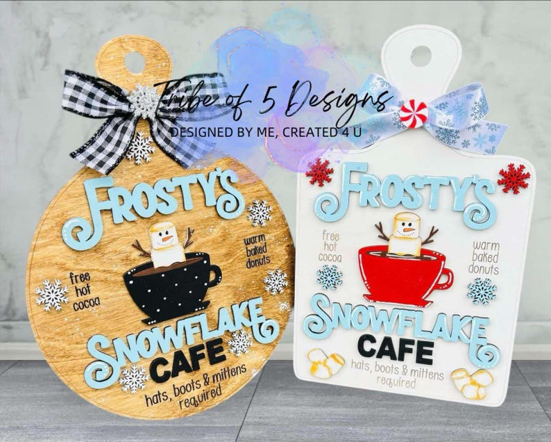 May include: Two wooden signs with a winter theme. Both signs are round and feature the text "Frosty's Snowflake Cafe" with a snowman and a cup of hot cocoa. One sign is brown and the other is white.