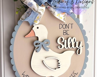 May include: Wooden door hanger featuring a white duck with a light blue bow, peach beak and feet. The sign reads "DON'T BE Silly, WE'RE GLAD YOU'RE HERE." A floral and denim bow tops the sign, adding a decorative touch.