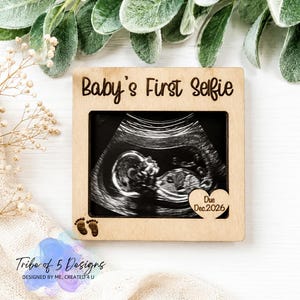 May include: A square light-colored wooden frame featuring the engraved text Baby's First Selfie at the top, two small footprints in the bottom left corner, and a heart-shaped cutout in the bottom right with the engraved text Due Dec. 2026.