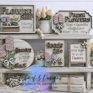 May include: A rustic wooden display featuring flower shop signs and decorative floral accents. The signs include text like "Fresh Flowers" and "Seeds 25¢". The display also showcases a vase of tulips and hyacinths.