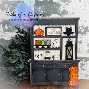 May include: A black and white Halloween-themed cabinet with decorative elements. It features a pumpkin, a bat, the word "BOO", a ghost, and the text "Happy Halloween". The cabinet has a lantern and stacked pumpkins. The text "Tribe of 5 Designs" is visible.