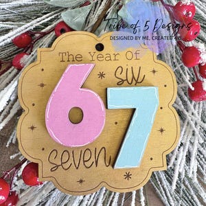Peut inclure: Ornement en bois avec le texte "The Year Of Six" et les chiffres "67" en rose et bleu. Le mot "seven" est également présent. L'ornement est décoré de petits accents d'étoiles et entouré de verdure festive et de baies rouges.
