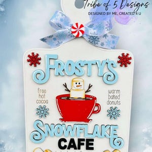 May include: A white wooden sign with a blue and white snowflake design. The sign says "Frosty's Snowflake Cafe" with a snowman holding a cup of hot cocoa. The sign also says "free hot cocoa" and "warm baked donuts".