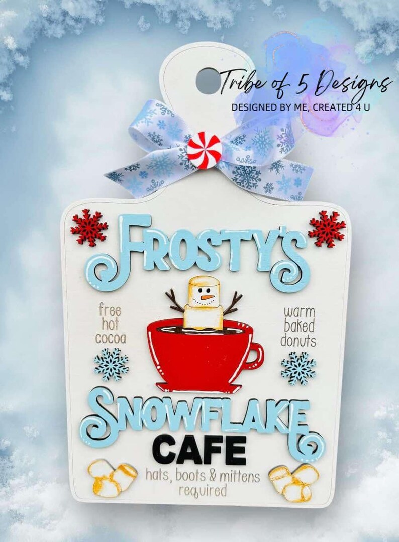 May include: A white wooden sign with a blue and white snowflake design. The sign says "Frosty's Snowflake Cafe" with a snowman holding a cup of hot cocoa. The sign also says "free hot cocoa" and "warm baked donuts".