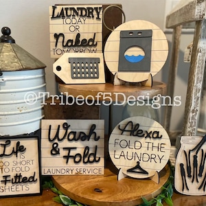 May include: A selection of wooden laundry signs with black lettering. Signs feature phrases such as "Laundry Today or Naked Tomorrow," "Wash & Fold Laundry Service," and "Life is Too Short to Fold Fitted Sheets."