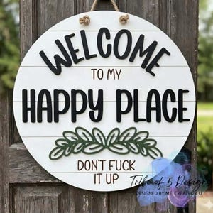 May include: A circular wooden sign, painted white, with the words "WELCOME TO MY HAPPY PLACE" in black. Below, it reads "DON'T FUCK IT UP". The sign features a green leaf design and is suspended by a rope.