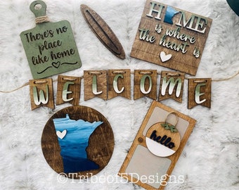 May include: A collection of wooden home decor items, including a cutting board with the text "There's no place like home", a "WELCOME" banner, and a sign that reads "HOME is where the heart is". The items are in shades of brown, green, and white.