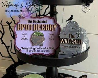 以下が含まれることがあります： ハロウィーンをテーマにした3段トレイ。装飾用の木製サインが飾られています。サインには、お化け屋敷、薬局の看板、墓石、幽霊、「Hush」と書かれた看板などがあります。トレイは黒いネットとカボチャで飾られています。