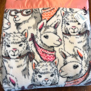 May include: A white fleece blanket with a pink satin border features a repeating pattern of llamas wearing colorful scarves. The blanket has a tag that reads "60x65" and "Northern Blessings Fleece Blanket".