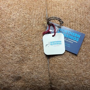 May include: A large bundle of light brown alpaca fiber. A tag with the words "Northern Blessings" and a logo of an alpaca is attached with a string. The fiber appears soft and fluffy, suitable for crafting.