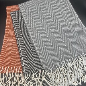 Puede incluir: Tres bufandas con un patrón de espiga. Las bufandas son en tonos de gris, negro y naranja. Las bufandas tienen flecos blancos.