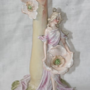 May include: A porcelain figurine featuring a figure standing beside a tall, pale yellow cylindrical structure. The figure wears a flowing, light pink and white garment and holds a large, pale pink flower. Another flower is attached to the column.