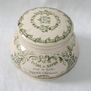 Op de afbeelding: Een ronde, crèmekleurige keramische pot met bijpassend deksel, voorzien van groene decoratieve krullen en bloemmotieven. De tekst op de voorzijde luidt CRÈME de SAVON pour la barbe. ROGER & GALLET. PARIS FRANCE, met een monogram op het deksel.