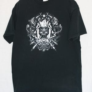Op de afbeelding: Zwart t-shirt met een gedetailleerde, grijswaardenafbeelding. Het ontwerp toont een demonisch gezicht, omringd door gestileerde figuren en abstracte elementen. Het t-shirt hangt aan een zwarte hanger.