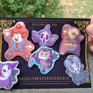 May include: Set of six handmade enamel pins featuring various cartoon characters. The pins are colourful and feature a variety of designs, including a clown, a ghost, a creature with a knife, a creature with a mask, a creature with a hat, and a creature with a skull. The pins are displayed on a black background with gold trim and the text "HANDMADE PIN" and "DESIGNMAIDENSTUDUIOS".