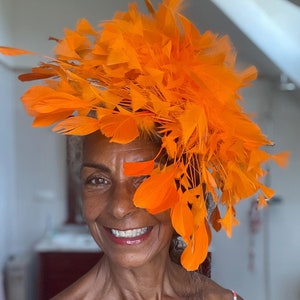Könnte beinhalten: Eine Frau trägt einen leuchtend orangefarbenen Federhut. Der Hut ist groß und bedeckt den größten Teil ihres Kopfes. Sie lächelt und schaut in die Kamera.
