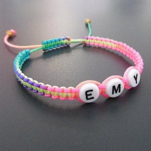 May include: A multi-colored friendship bracelet with a pink, green, and purple cord. The bracelet has three white beads with the letters "EMY" spelled out.