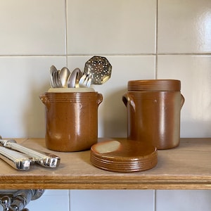 Puede incluir: Dos botes de cocina de cerámica marrón con asas, uno con cubiertos y una cuchara ranurada. También hay una pila de posavasos de cerámica marrón y un tercer bote. Los artículos están sobre una estantería de madera.