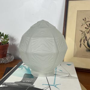 Op de afbeelding: Een bolvormige glazen lampenkap met een matte afwerking en een geometrisch reliëfpatroon met ruit- en driehoeksvormen. De kap heeft een ronde opening aan de onderkant en een gefacetteerde bovenkant met een textuur van herhalende lijnen.