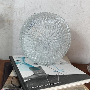 May include: A clear, faceted glass bowl with a starburst design. The bowl is spherical and rests on a stack of books, one of which has the text "NEUZEITLIC MODERN L" and "ARNOLDSCHE ART PUBLISHERS".