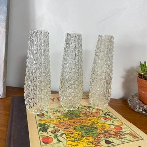 Pode incluir: Três vasos de vidro transparente com um padrão geométrico texturizado em forma de diamante no exterior. Cada vaso tem uma forma cilíndrica cónica com uma abertura estreita no topo e uma base plana, dispostos em fila.