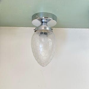 Op de afbeelding: Een chromen plafondlamp met een matglazen kap. De kap heeft een druppelvorm met een honingraatpatroon. De lamp is gemonteerd op een wit plafond.