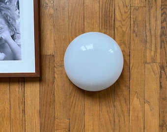 May include: A white, spherical light fixture is centered on a wooden floor. The fixture has a smooth, opaque surface. A framed black and white photograph is partially visible in the upper left corner. The wooden floor has a warm, natural wood grain.