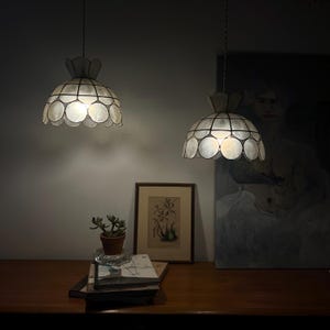 Op de afbeelding: Twee koepelvormige hanglampen gemaakt van doorschijnende, ronde schelpsegmenten gerangschikt in een geschulpt patroon. Elke lamp heeft een donker metalen frame dat de overlappende schelpschijven vasthoudt en een zacht licht uitstraalt.