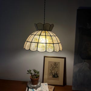 Könnte beinhalten: Eine Hängelampe mit einem gewellten, kuppelförmigen Schirm aus durchscheinenden Paneelen. Der Schirm zeigt ein Gittermuster mit handgemalten gelben Blumen und grünen Blättern. Die Lampe ist an einem gedrehten Kabel befestigt und leuchtet warm.