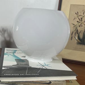May include: A spherical white glass lamp shade with a smooth, opaque finish and a circular opening at the base. The glass has a glossy surface that reflects light, creating a bright, clean appearance. The shade features a simple, rounded form.