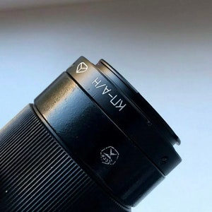 Può includere: Un obiettivo per fotocamera nero con la scritta "KF-4H" stampata sul lato.