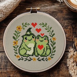 Może przedstawiać: Wzór haftu krzyżykowego w drewnianym tamborku przedstawia dwie zielone żaby z sercami, otoczone kwiatami i zielenią. Jedna żaba ma na sobie kapelusz czarownicy. Tekst "CROSS STITCH PATTERN PDF" znajduje się w prawym dolnym rogu.