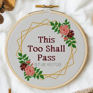May include: A circular wooden embroidery hoop containing a cross-stitch design with the text This Too Shall Pass and But Like Holy Flick in dark red, surrounded by a geometric gold frame and floral accents in shades of pink and dark red.