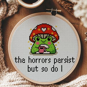 May include: A cross-stitch design in a wooden hoop shows a green frog under a red mushroom, holding a pink cup. The text reads "the horrors persist but so do I." A cup of coffee and a beige scarf are in the background.